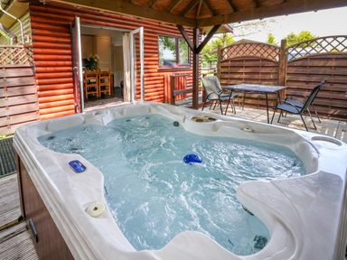 An outdoor hot tub on a wooden deck under a roof next to a wooden cabin with open doors and a table with chairs at The Blacktail in Llanerch Park near St Asaph