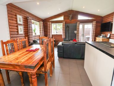 A dining table with six wooden chairs and a living area with black sofas a wood burning stove and a television in a log cabin at The Buck Llanerch Park near St Asaph
