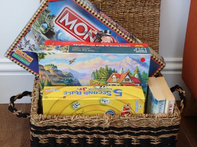 A woven basket containing board games and two paperback books on a wooden floor
