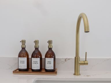 Three brown pump bottles labeled hand wash hand lotion and dish soap on a wooden tray next to a brass kitchen faucet on a white marble countertop at Victory View in Pendeen