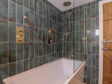 A bathtub with a glass partition in a tiled bathroom with brass fixtures and a wooden door at Victory View in Pendeen