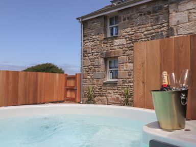 An outdoor hot tub with bubbling water beside a stone building and wooden fence with a champagne bucket and glasses at Victory View in Pendeen