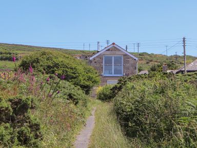 A stone house at the end of a narrow path with bushes and wildflowers on both sides at Victory View in Pendeen