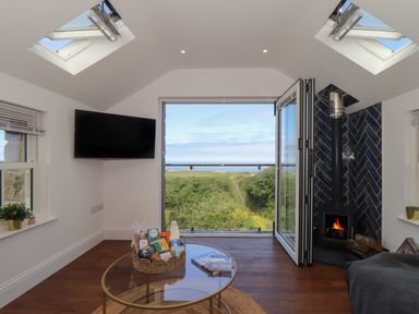 A living room with a glass coffee table basket with groceries a wall-mounted television a fireplace with black tiles and an open door showing a green landscape and sea view at Victory View in Pendeen