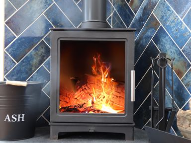 A wood burning stove with fire inside a room with blue tiled walls and a black ash bucket and fireplace tools nearby at Victory View Pendeen
