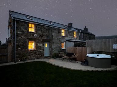 A stone house with lit windows a patio with chairs and a hot tub under a starry sky at Victory View in Pendeen