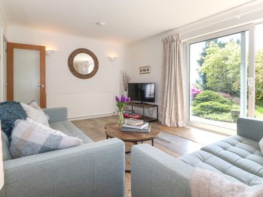 A living room with two sofas a round wooden coffee table a television on a stand and large glass sliding doors showing a garden at Sweden Bridge Cottage in Ambleside