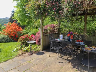 A garden patio with a metal dining table and chairs under a wooden pergola with flowers and food on the table at Sweden Bridge Cottage in Ambleside
