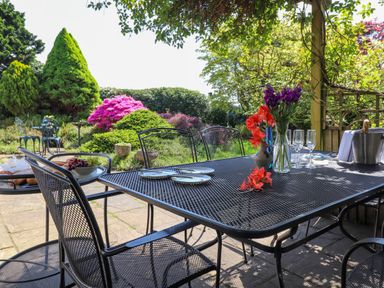 An outdoor dining area with a metal table and chairs flowers on the table and a garden with bushes and trees Sweden Bridge Cottage Ambleside