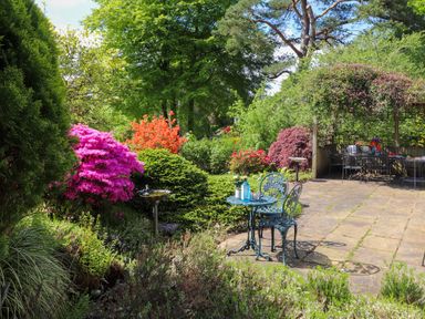 A garden with pink and orange flowering bushes a small metal table with chairs and a pergola covered seating area at Sweden Bridge Cottage Ambleside