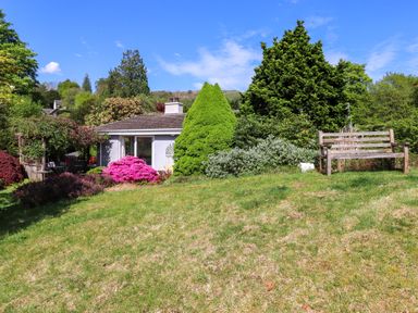 A garden with a wooden bench green bushes pink flowers and a house in the background at Sweden Bridge Cottage in Ambleside