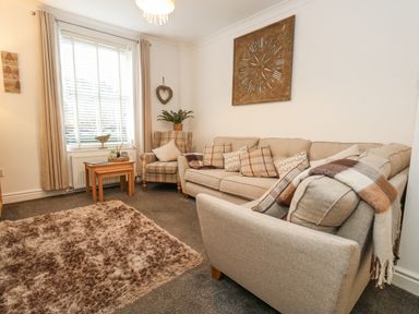 A living room with a beige sofa and armchair a wooden side table underneath a window with curtains and a wall clock at Garibaldi Cottage in Weymouth