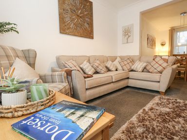 A living room with a beige corner sofa and an armchair with cushions a wooden table with plants and a book and a clock on the wall at Garibaldi Cottage in Weymouth
