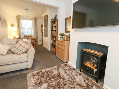 A living room with a sofa and cushions a television above a fireplace and a dining area with a table and chairs at Garibaldi Cottage in Weymouth