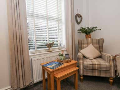 A small sitting area with a plaid armchair a cushion nested tables with plants and a book near a window with blinds and curtains at Garibaldi Cottage in Weymouth