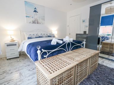 A bedroom with a white metal bed frame blue bedding and rolled towels on the bed bedside tables with lamps a wooden storage bench a grey dresser and a mirrored wardrobe at Garibaldi Cottage in Weymouth