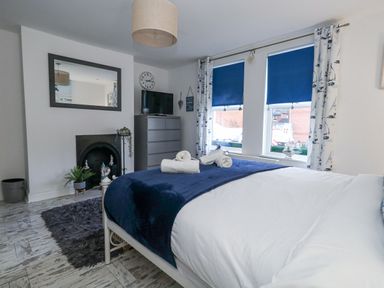A bedroom with a bed, blue and white bedding, a dresser with a television, a fireplace, two windows with blue blinds, and patterned curtains at Garibaldi Cottage in Weymouth