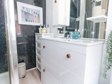 A bathroom with a white sink cabinet a small white drawer unit a soap dispenser and lotion bottles near a shower at Garibaldi Cottage in Weymouth