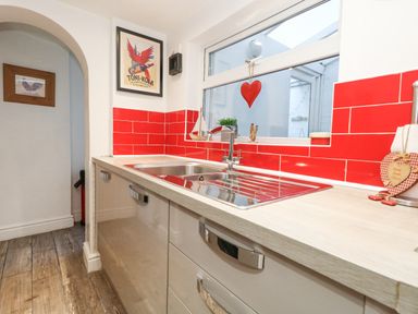 A kitchen countertop with a stainless steel sink red tile backsplash and window above with decorative items at Garibaldi Cottage in Weymouth