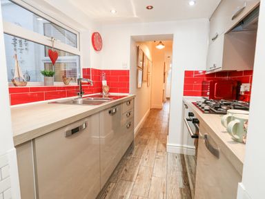 A galley kitchen with beige cabinets red tiled backsplash a stainless steel sink and gas stove at Garibaldi Cottage in Weymouth