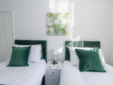 A bedroom with two single beds green pillows and a white nightstand with a lamp and coasters at Garibaldi Cottage in Weymouth