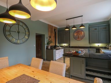 A kitchen and dining area with a wooden table, upholstered chairs, hanging lights, a large wall clock, and green cabinetry at Llwynderw Hall in Abergwesyn near Llanwrtyd Wells