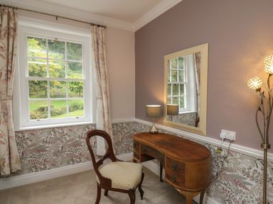 A room with a wooden desk an upholstered chair a table lamp a large mirror floral curtains and patterned wallpaper at Llwynderw Hall in Abergwesyn near Llanwrtyd Wells