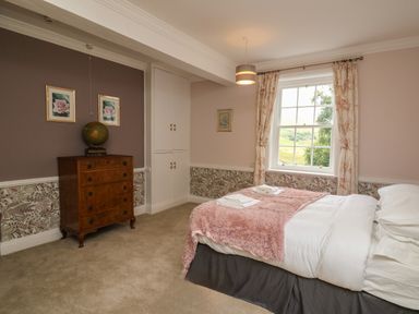 A bedroom with a bed covered with a pink throw and white linens a wooden chest of drawers and a window with floral curtains at Llwynderw Hall in Abergwesyn near Llanwrtyd Wells