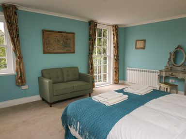 A bedroom with a bed covered in white and blue linens a green sofa two windows with patterned curtains and a vanity with a mirror at Llwynderw Hall in Abergwesyn near Llanwrtyd Wells