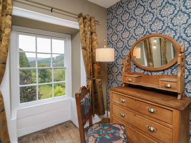 A wooden dresser with an oval mirror a chair with floral upholstery a floor lamp and a window with yellow curtains overlooking greenery at Llwynderw Hall in Abergwesyn near Llanwrtyd Wells