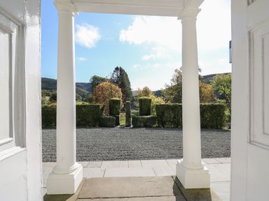 View from a doorway with white columns onto a gravel pathway with trimmed hedges and trees in the background at Llwynderw Hall in Abergwesyn near Llanwrtyd Wells