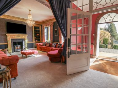 A living room with red sofas a fireplace chandelier and an open door leading outside at Llwynderw Hall in Abergwesyn near Llanwrtyd Wells