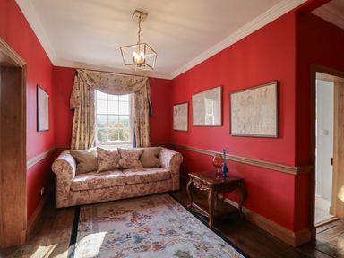 A sitting room with a patterned sofa a window with curtains framed maps on red walls and a wooden side table at Llwynderw Hall in Abergwesyn near Llanwrtyd Wells