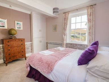 A bedroom with a bed with purple pillows and a pink blanket a wooden chest of drawers and a window with floral curtains at Llwynderw Hall in Abergwesyn near Llanwrtyd Wells