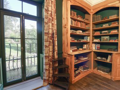 A corner bookshelf filled with books next to a glass door with curtains and a wooden spiral staircase at Llwynderw Hall in Abergwesyn near Llanwrtyd Wells