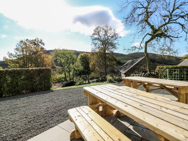 An outdoor area with wooden picnic tables and benches overlooking trees hills and a stone building at Llwynderw Hall in Abergwesyn near Llanwrtyd Wells