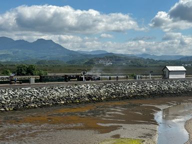 A train on a track with mountains in the background and a small white building near a rocky shoreline at Estuary Escape in Porthmadog