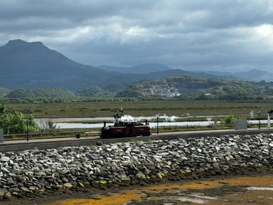 A steam train on a railway track near a rocky shoreline with mountains and cloudy sky in the background at Estuary Escape in Porthmadog
