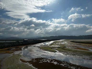 A tidal river with exposed mudflats and small buildings on a stone embankment under a cloudy sky at Estuary Escape in Porthmadog
