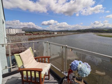 A balcony with a green chair and a table with flowers and drinks overlooking a river and a train on a track at Estuary Escape in Porthmadog