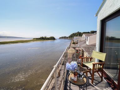 A balcony with a chair small table with flowers and a wine bottle overlooking a river and houses along the shore at Estuary Escape in Porthmadog