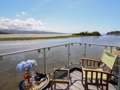 A balcony with a chair a small table holding a bottle flowers glasses and binoculars overlooking a river and landscape at Estuary Escape in Porthmadog