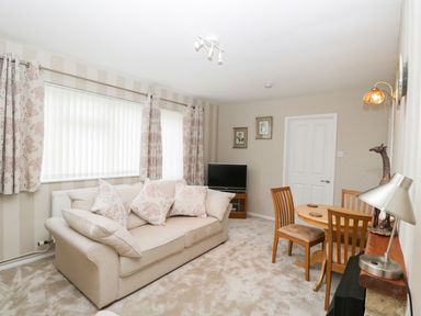 A living room with a beige sofa and floral cushions a small dining table with four chairs a television and floral curtains at Ground Floor Annex in Milton-Under-Wychwood