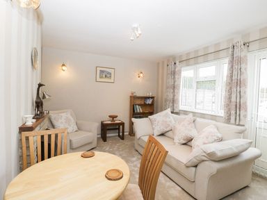 A living room with a round wooden table, beige sofas, patterned cushions, curtains, and a small wooden bookcase at Ground Floor Annex in Milton-Under-Wychwood