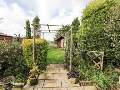 A garden with a wooden pergola pathway leading to a small wooden shed surrounded by green bushes and trees at Ground Floor Annex in Milton-Under-Wychwood