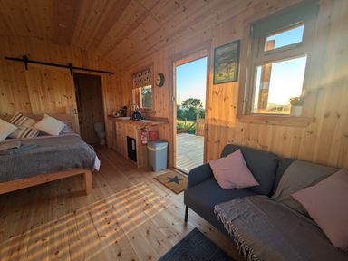 A wooden interior room with a bed sofa kitchen area and a door leading outside at Hafan Gelli Sheperds Hut - Tudwal in Pwllheli
