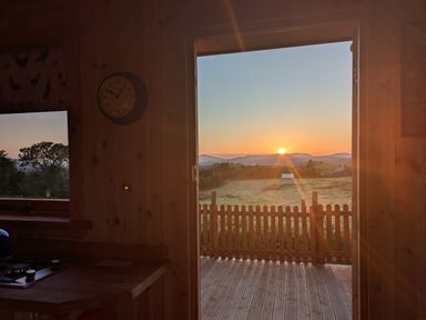 A wooden kitchen interior with a clock on the wall and an open door leading to a deck overlooking a sunrise and fields at Hafan Gelli Sheperds Hut - Tudwal in Pwllheli