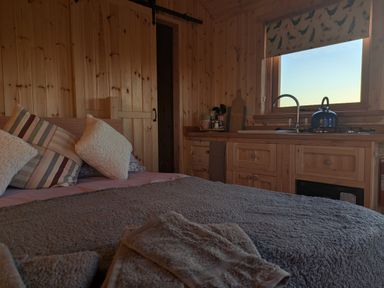 A wooden interior room with a bed with cushions and a kitchen area with cabinets and a window at Hafan Gelli Sheperds Hut - Tudwal in Pwllheli