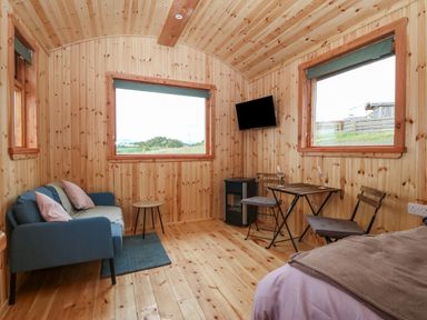 A wooden interior room with a blue sofa and pillows a small round table a wall-mounted television a small dining table with two chairs and a bed Hafan Gelli Sheperds Hut - Tudwal Pwllheli