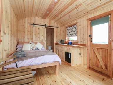 An interior of a wooden room with a bed, kitchen area, wooden door, and a sliding barn door at Hafan Gelli Sheperds Hut - Tudwal in Pwllheli